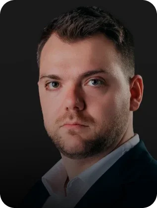 Professional headshot of a man wearing a dark suit jacket and white shirt against a black background.